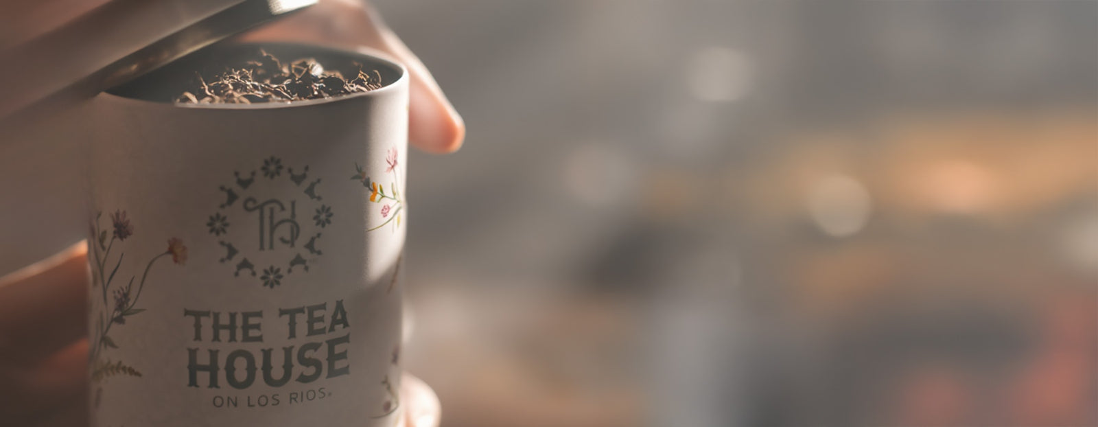 White tea tin with 'The Tea House' branding filled with tea leaves, held by a hand against a blurred background.