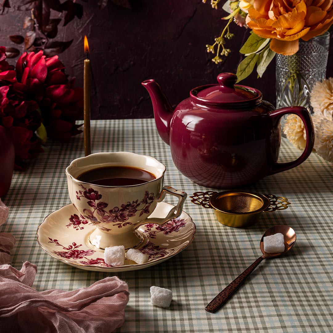 Tea set with a teapot, cup, and saucer on a checkered tablecloth with flowers and a candle in the background.