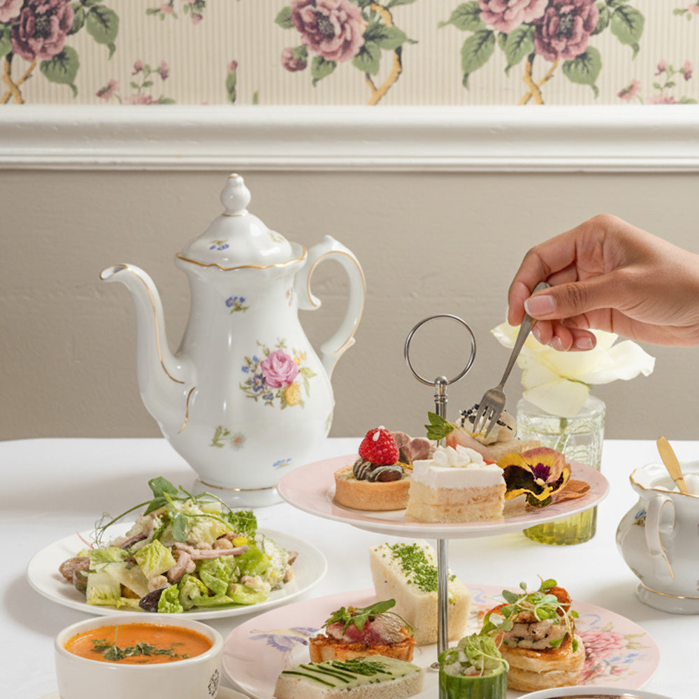 Tea party setup with teapot, sandwiches, and salads on a floral wallpapered wall background.