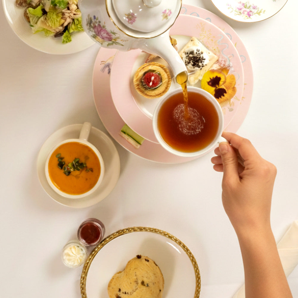 Tea cup with a hand pouring tea, surrounded by various dishes on a white table.
