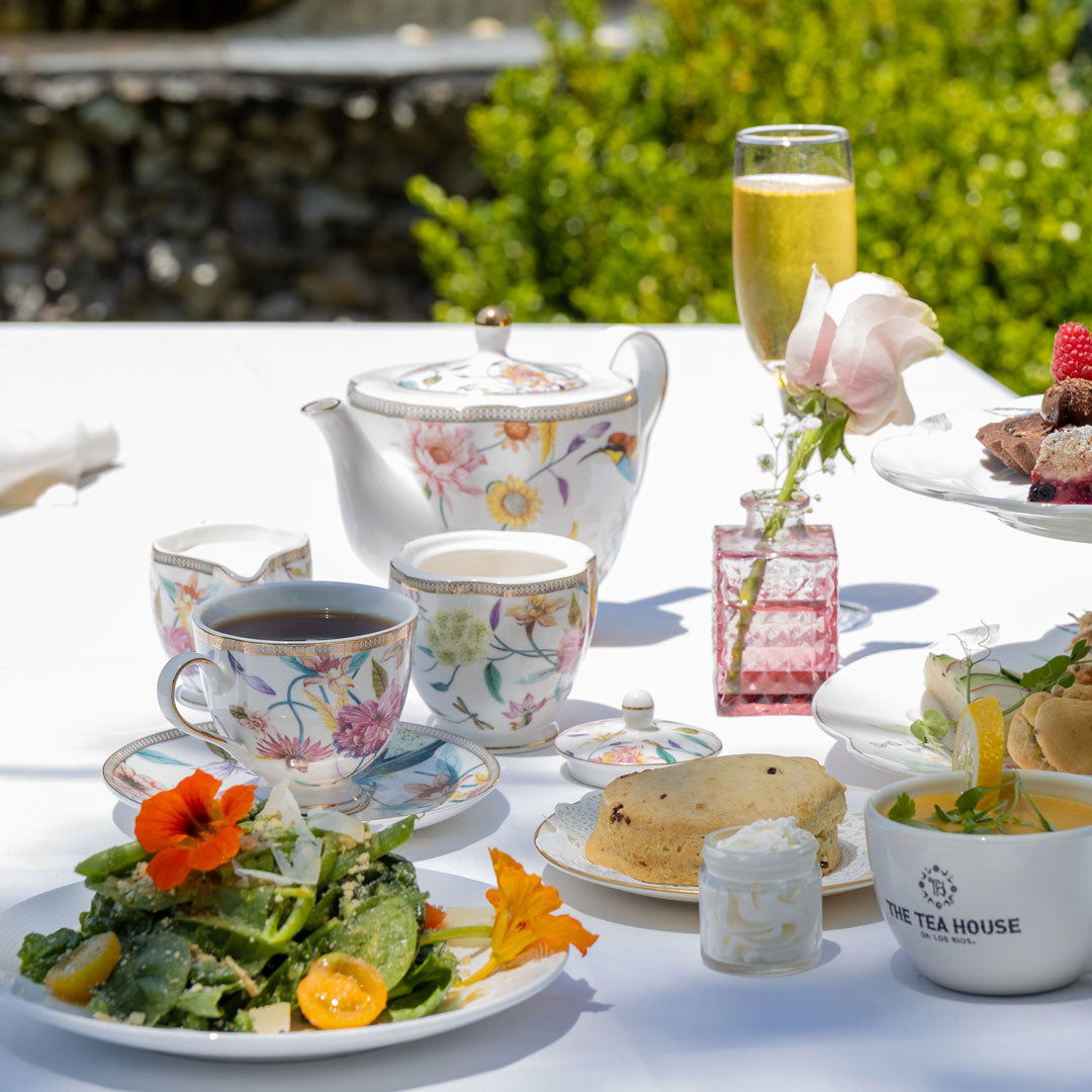 Tea set with a teapot, cups, and plates of food on a table outdoors.