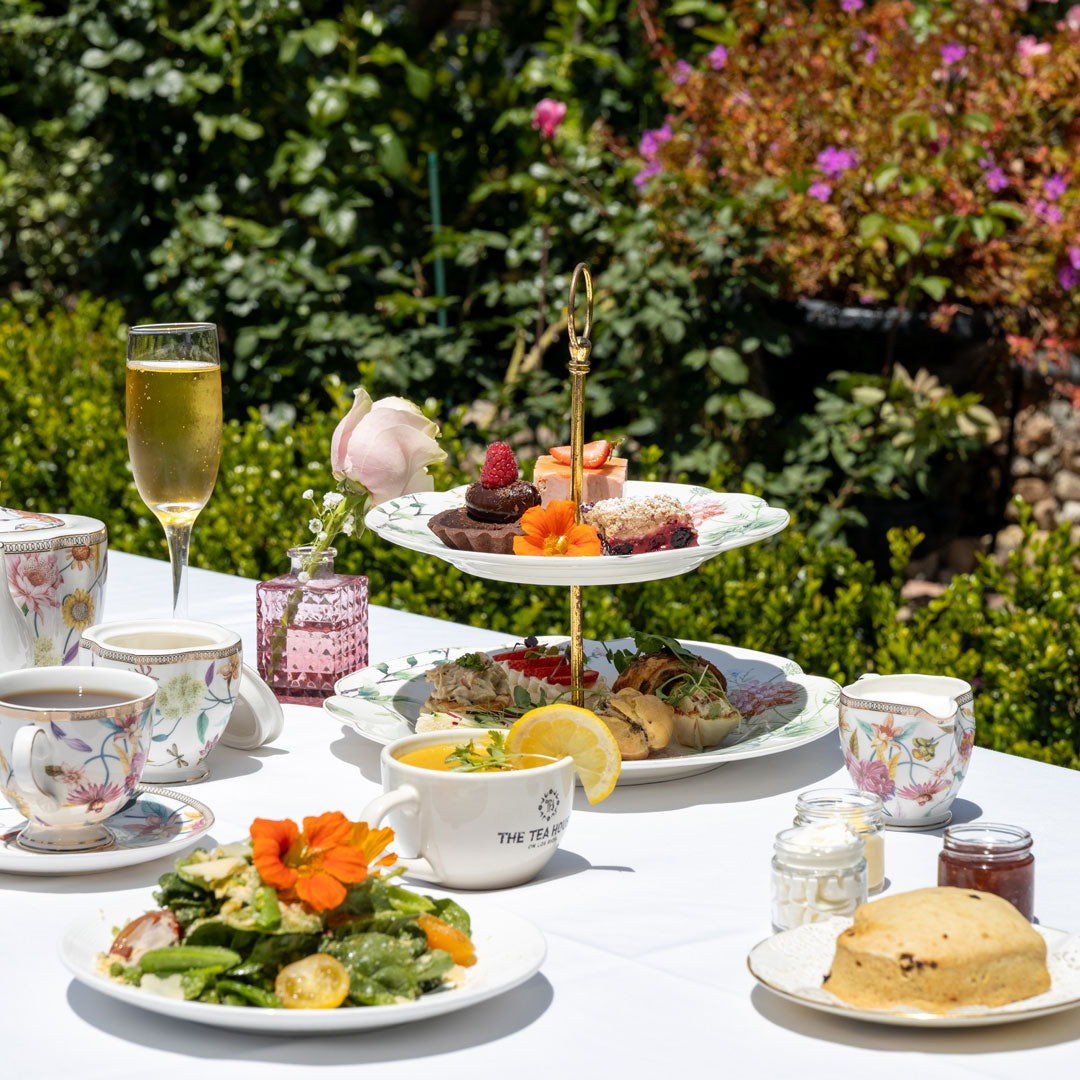 Table set for afternoon tea with a three-tiered cake stand, teacups, and assorted treats outdoors.