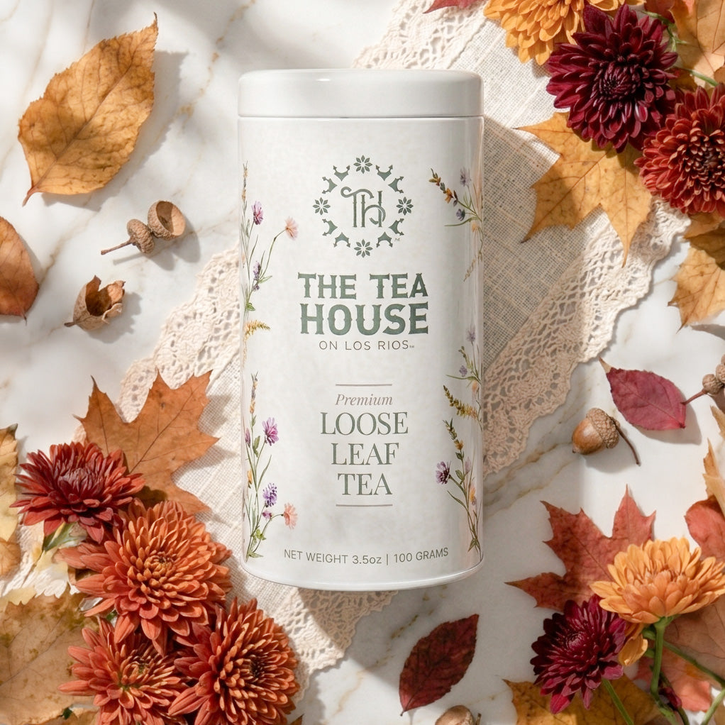 A white cylindrical tin of The Tea House on Los Rios Premium Loose Leaf Tea is styled in an autumn flat-lay with dried leaves, acorns, lace, and orange-red flowers on a white marble surface.
