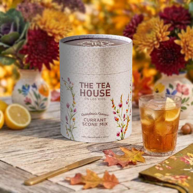 A cylindrical tin of The Tea House On Los Rios Currant Scone Mix stands on a rustic wooden table next to a glass of iced tea with lemon slices. The background is a soft-focus autumnal setting with flowers and fall leaves.
