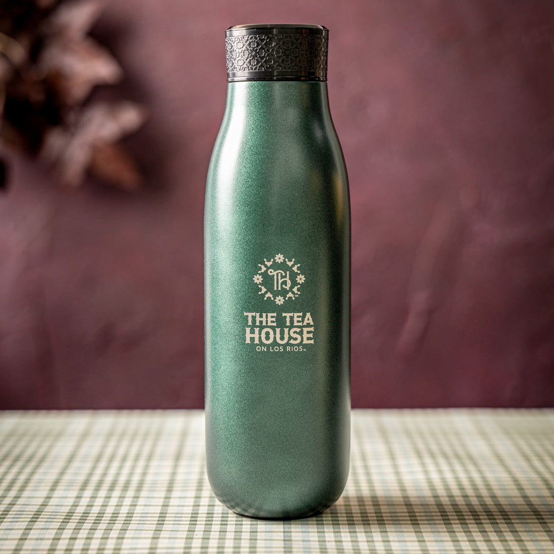 A matte dark green insulated water bottle with a black textured lid, standing on a green-and-white checkered tablecloth against a dark mauve background. The bottle features a gold logo for 'THE TEA HOUSE ON LOS RIOS'.