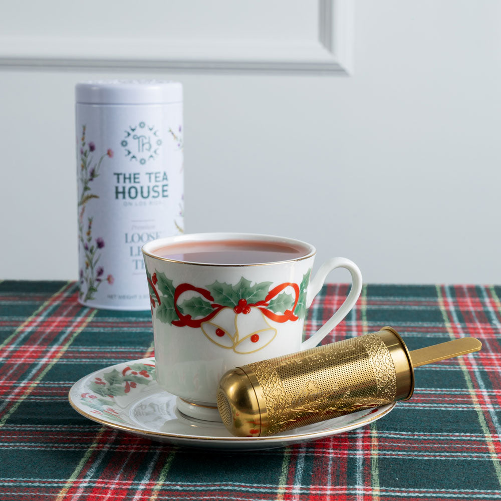 Tea cup with festive design on a plaid tablecloth, next to a tea canister and gold tea infuser.