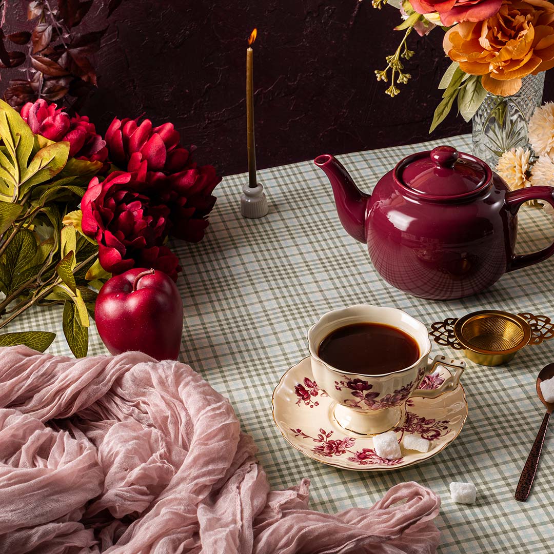 Tea set with a teapot, cup, and saucer on a checkered tablecloth with flowers and a candle.
