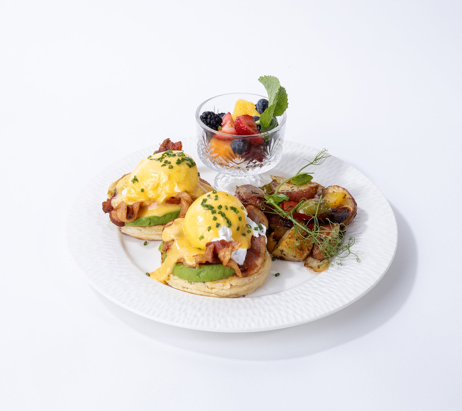 A plated breakfast food presentation on a white plate, featuring two eggs Benedict with avocado and bacon, a side of roasted potatoes, and a small glass bowl of fresh fruit and berries, set against a clean white background.