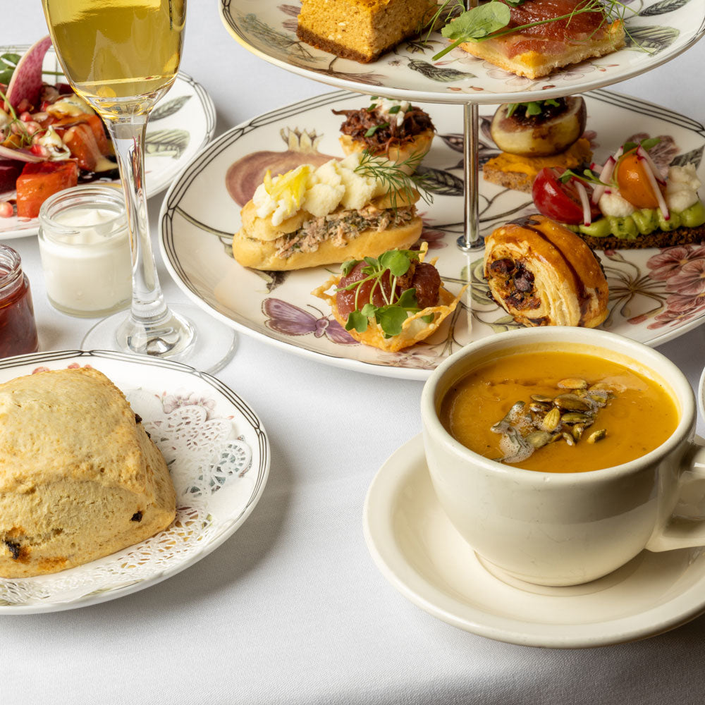 An elegant afternoon tea spread on a white tablecloth, featuring a three-tiered floral stand with sandwiches, pastries, and fruit, alongside a bowl of orange soup, a scone, a glass of white wine, and small jars on a clean white background.