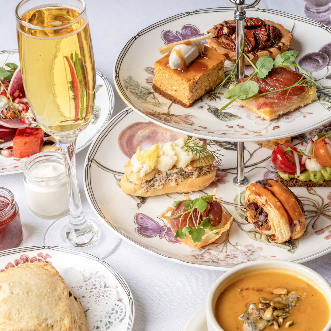 An elegant two-tiered floral serving platter filled with assorted gourmet afternoon tea, alongside a filled champagne flute, a bowl of pumpkin soup, and other tea-time dishes on a white tablecloth.