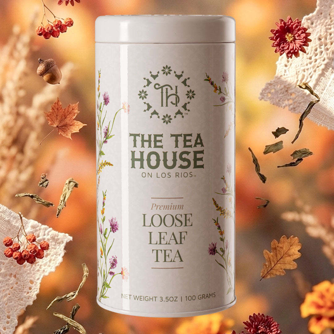 A white cylindrical tin of The Tea House On Los Rios Premium Loose Leaf Tea, surrounded by autumn-themed decorative elements like dried leaves, lace, and berries on a warm, blurred background.