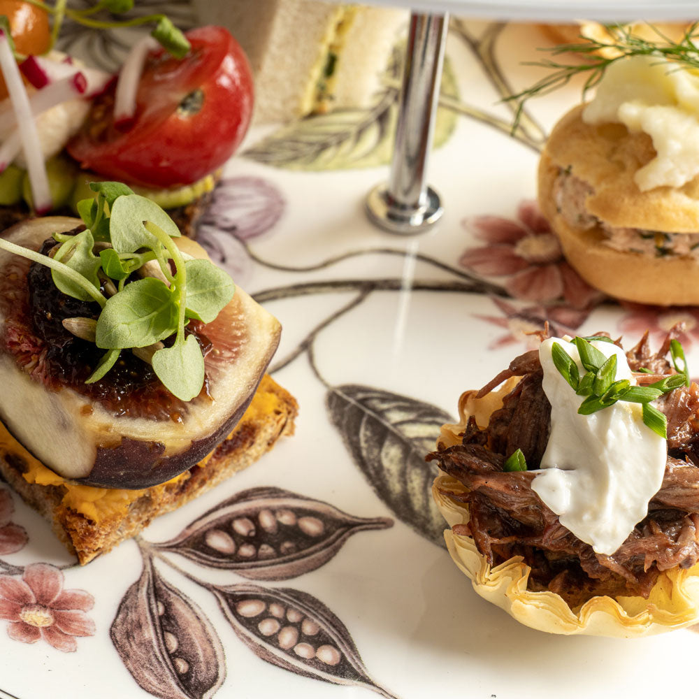 Close-up of plated gourmet sandwiches and small savory pastries on a floral ceramic plate, with a metal stand leg visible in the background.
