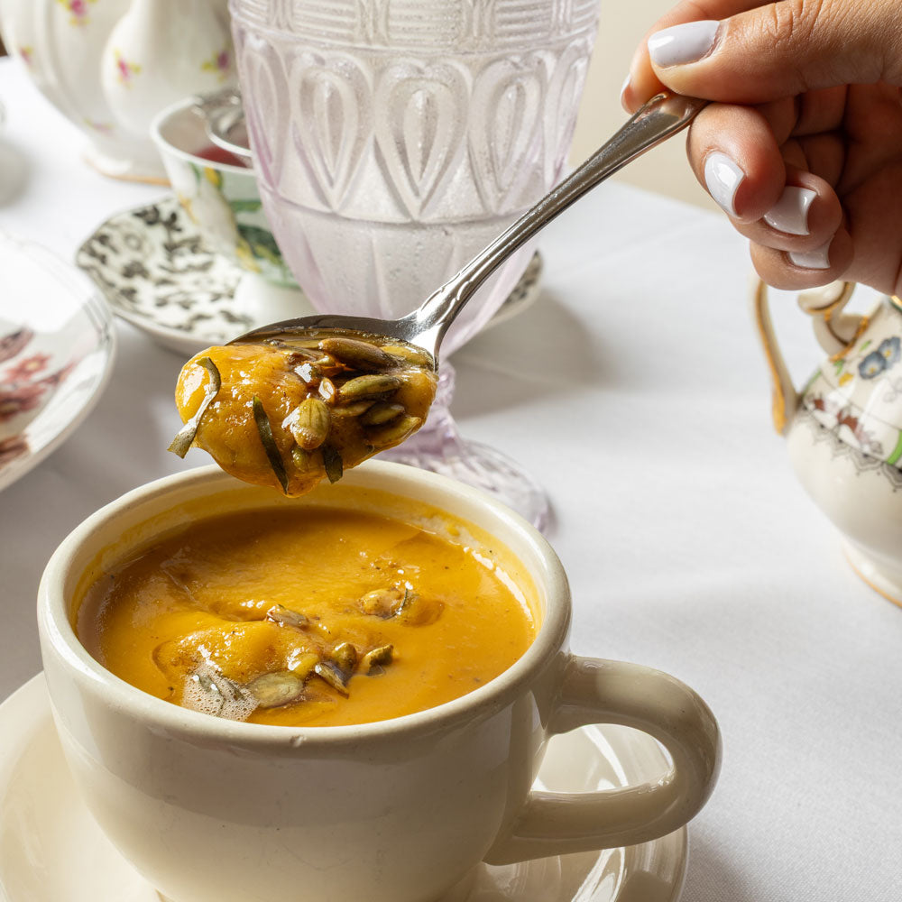 A close-up of a white ceramic teacup filled with orange-yellow soup, with a hand holding a spoonful of the soup above the cup on a white tablecloth. Teaeware and a glass are softly blurred in the background.