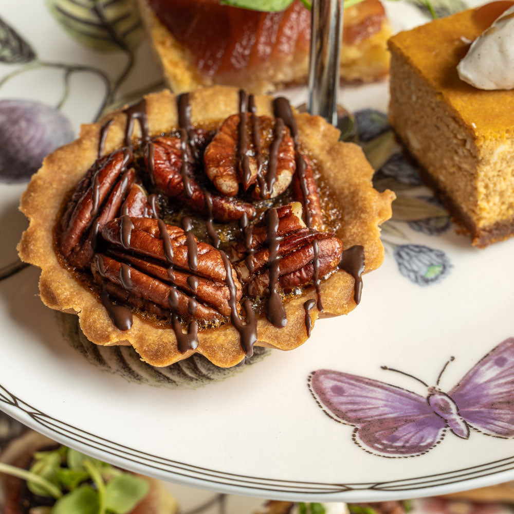 A close-up of a pecan tart on a white plate with a purple butterfly print, alongside other desserts including a slice of pumpkin pie and a brownie.
