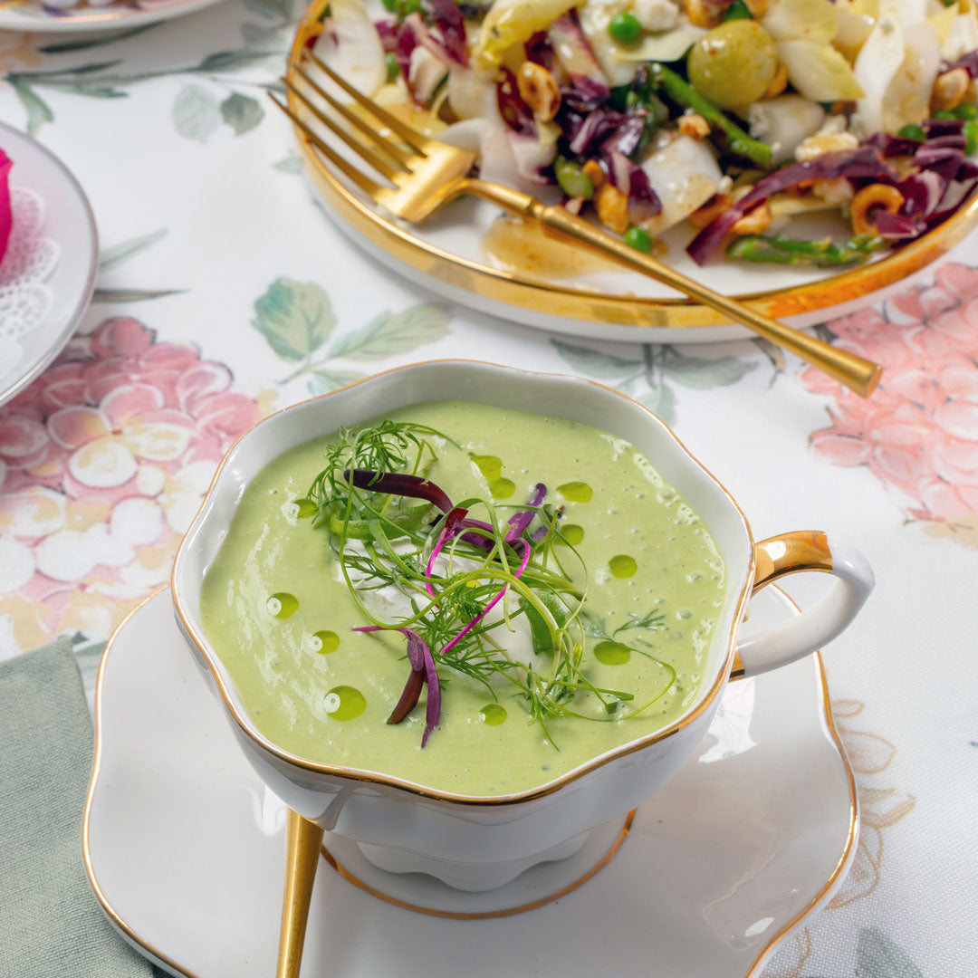 Green soup in a decorative bowl on a floral tablecloth with a salad in the background