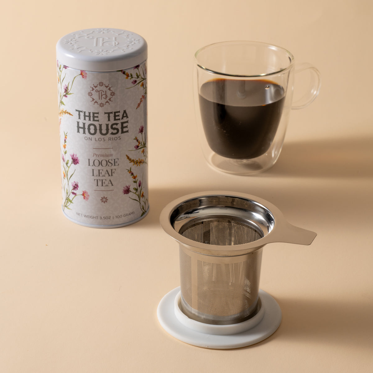 Tea house tea tin, glass with tea, and metal tea infuser on a beige background